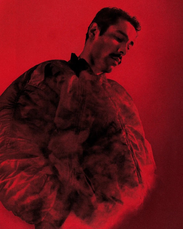 M Le Maudit in a puffer jacket surrounded by smoke, red-toned portrait
