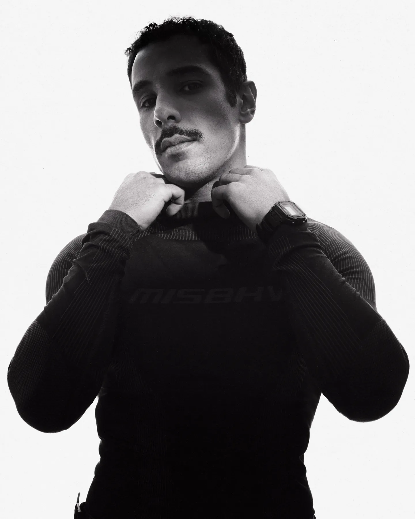 M Le Maudit in a black MISBHV top, hands at his collar, on a white background