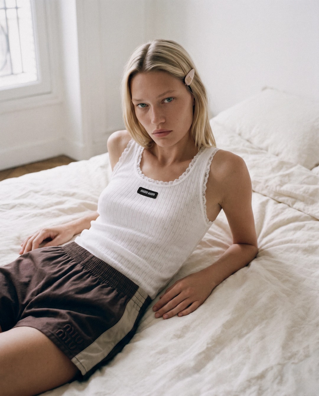 Miu Miu x New Balance AI editorial: model in a white Miu Miu tank top and brown shorts sitting on an unmade bed