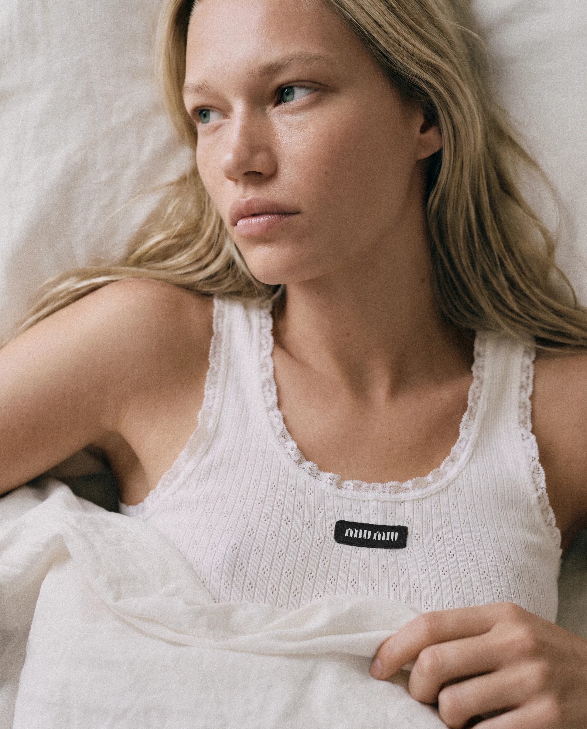 Close-up of the model lying on white sheets, Miu Miu logo on the tank top