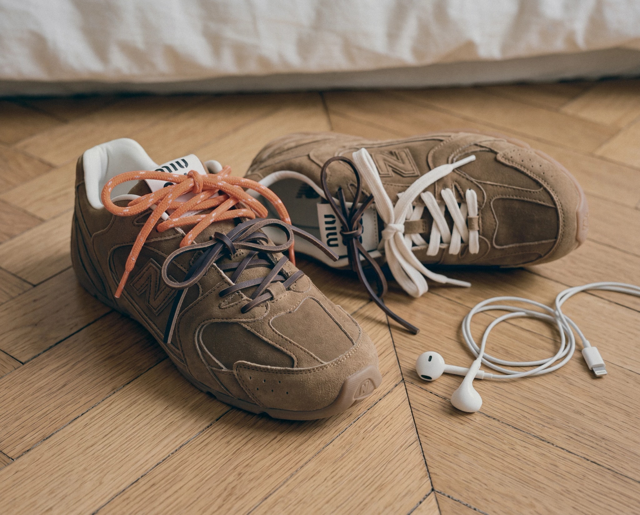 Pair of Miu Miu x New Balance sneakers on a parquet floor next to wired earphones